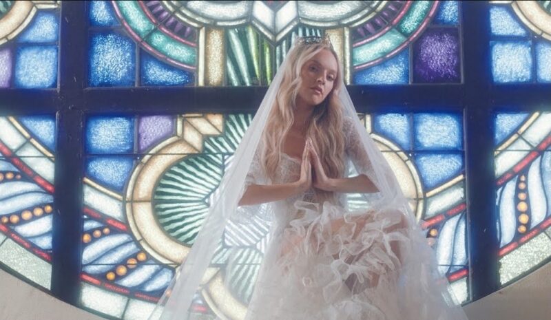 Uma mulher branca e loira com um vestido branco, véu de noiva e uma coroa, está sentada em frente a um grande vitral colorido comum em igrejas da Idade Média. O vitral é arredondado e usa cores vibrantes como azul, verde e amarelo. A mulher está no centro da imagem com a cabeça levemente inclinada para o lado direito. Suas mãos estão unidas em posição de oração e sua expressão facial é séria. 