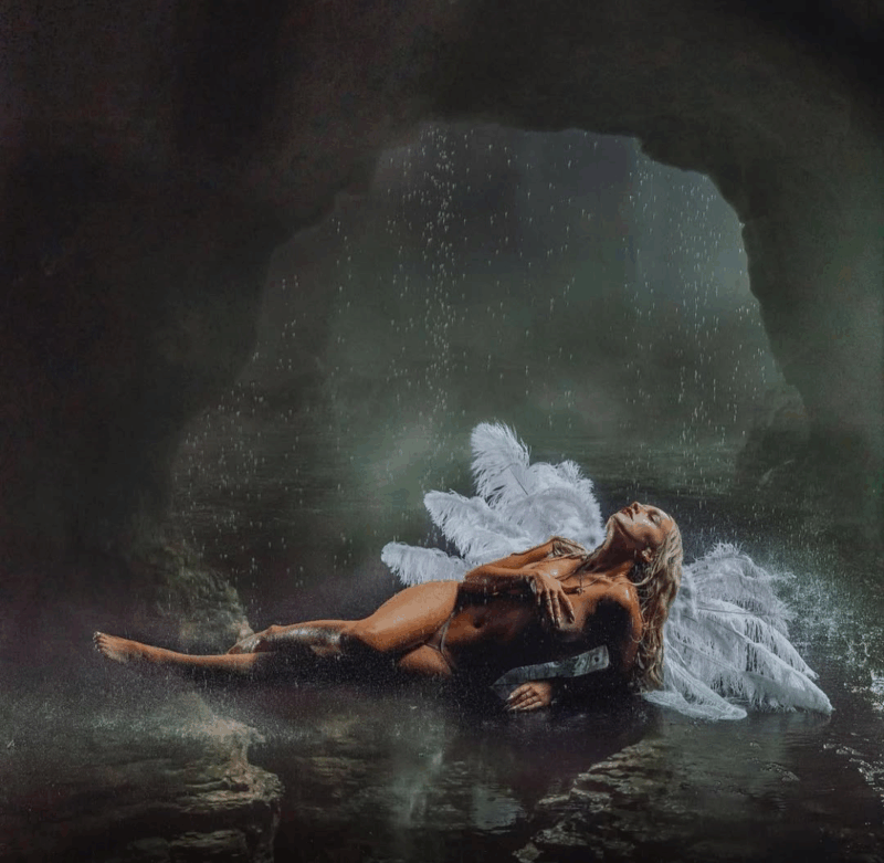 Uma mulher branca e loira está deitada em uma caverna escura e úmida, com uma chuva leve caindo ao redor dela. Ela está nua, com apenas as partes íntimas cobertas. Ela tem grandes asas brancas de penas nas costas, simbolizando um anjo caído, enquanto mantém uma expressão contemplativa e olhos fechados, apreciando a chuva.