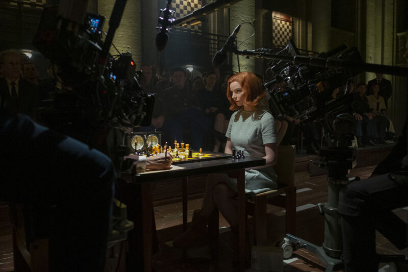 A foto mostra um set de filmagem escuro, com câmeras e microfones que cercam uma jovem mulher de cabelo curto ruivo sentada diante de um tabuleiro de xadrez iluminado, em um vestido cinza. Ao fundo, espectadores observam atentamente enquanto a equipe técnica registra cada detalhe da cena, criando um contraste entre o foco dramático da partida e a produção ao redor. 