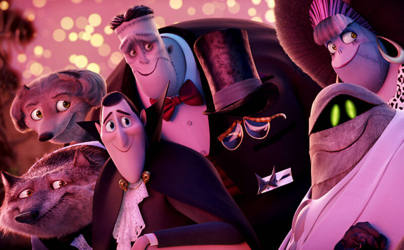 Cena de Hotel Transilvânia 2. A imagem mostra um grupo diverso de personagens de Hotel Transilvânia, todos reunidos de forma próxima e calorosa. O enquadramento fechado foca nos rostos e expressões, transmitindo união e companheirismo. Entre os personagens em destaque estão o Conde Drácula, em seu traje clássico e com expressão amigável; Frank, marcado pelos pontos característicos e olhar bondoso; além de um lobisomem, uma múmia e outras figuras icônicas da série. Cada um exibe traços físicos e expressões que reforçam suas personalidades, enriquecidos por detalhes de trajes e acessórios. O estilo em animação 3D valoriza cores vibrantes, texturas bem-feitas e iluminação suave, criando um ambiente aconchegante e visualmente atraente.