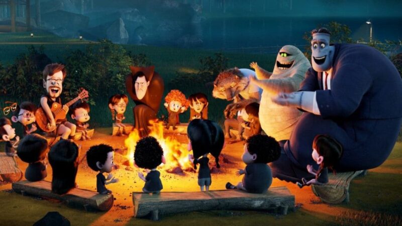 Cena de Hotel Transilvânia 2. A imagem mostra um grupo de personagens de animação reunidos ao redor de uma fogueira, em um momento de convívio noturno cheio de música e diversão. Entre eles estão figuras icônicas como o Conde Drácula, com seu ar imponente mas descontraído, e o Monstro de Frankenstein, com seu porte marcante e expressão amistosa. Outros seres antropomórficos completam o círculo, cada um com traços físicos distintos e expressões que variam entre alegria, concentração e curiosidade, reforçando suas personalidades singulares. O destaque vai para o personagem que toca violão, animando o grupo e servindo como centro da interação. A iluminação quente da fogueira valoriza os rostos e corpos dos personagens, dando profundidade às suas expressões e ressaltando detalhes de roupas e características físicas. O estilo em animação digital de alta qualidade combina cores vibrantes e sombras realistas, criando uma atmosfera acolhedora. O cenário de floresta, com céu estrelado ao fundo, funciona como pano de fundo para a proximidade e o espírito de união dos personagens.