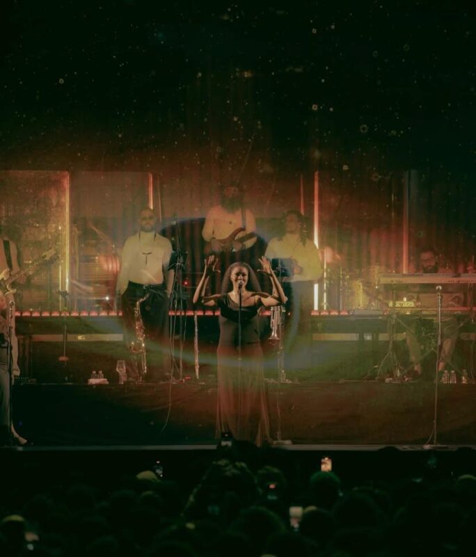 Na imagem, há uma mulher negra se apresentando no centro de um palco, em frente a um microfone com as duas mãos erguidas. Atrás dela, uma banda completa toca: músicos com saxofone, guitarra, teclado e percussão. Na frente do palco, é possível ver a silhueta da plateia, com várias pessoas registrando o momento com seus celulares. As luzes da imagem são quentes e difusas, criando um aspecto esfumaçado.  