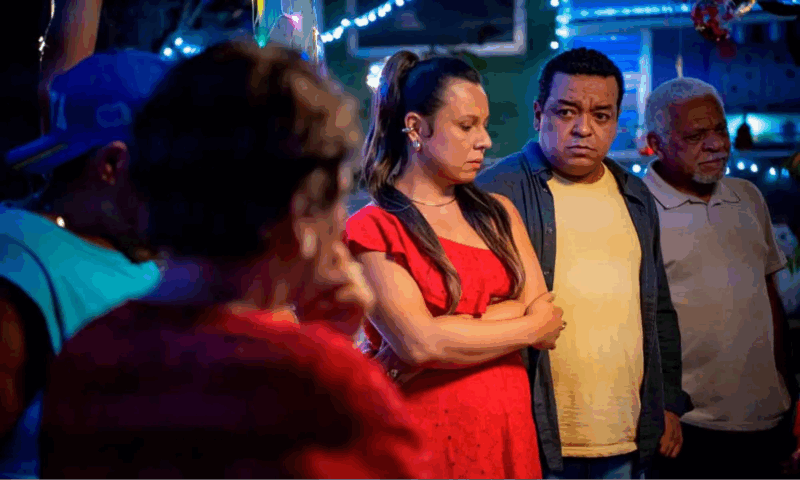 Quatro adultos negros estão de pé em uma área externa à noite, sob luzes pisca-pisca azuis e brancas desfocadas. Uma mulher de vestido vermelho no centro está de braços cruzados e expressão séria, olhando para a esquerda, assim como os dois homens ao seu lado. A tensão é visível em seus rostos.