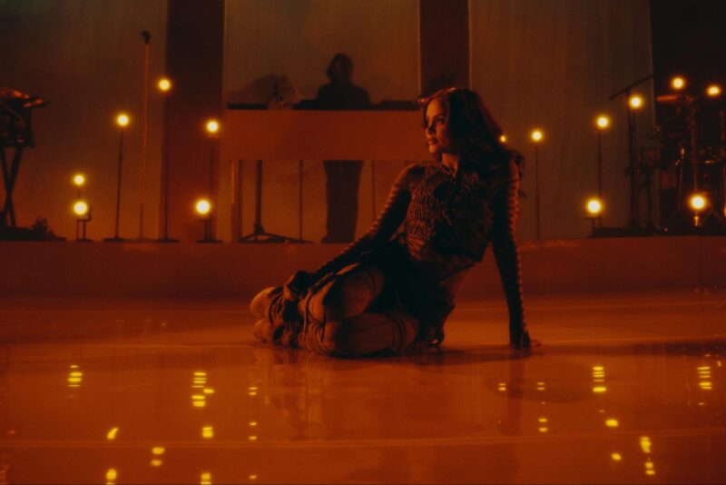 No centro da imagem, há uma mulher vestindo uma blusa e usando luvas e botas de cano alto. Ela está sentada de lado no chão de um palco enquanto olha para o lado esquerdo da foto. Ao fundo da imagem, é possível ver microfones, um teclado e partes de uma bateria. A iluminação da imagem é predominantemente laranja e amarela. 