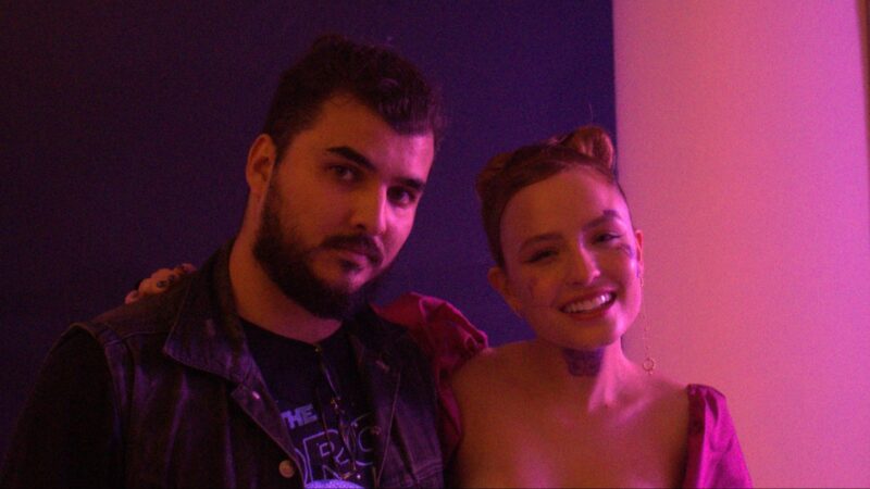 Na fotografia, duas pessoas posam lado a lado. A mulher à direita sorri com expressão leve, usando um vestido rosa com mangas bufantes e penteado elaborado e preso, enquanto o homem, branco de cabelo, barba e bigode negros, à esquerda mantém um olhar mais sereno, vestindo jaqueta jeans escura e blusa preta por baixo. Como cenário, tem-se uma iluminação rosa e arroxeada refletidas nas paredes.