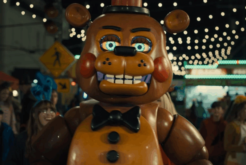 Cena de Five Nights at Freddy’s 2. A cena mostra um boneco do personagem Toy Freddy, do jogo "Five Nights at Freddy's", domina o centro da imagem. O boneco é marrom com detalhes em marrom claro e vermelho. Possui olhos grandes com íris verde-azuladas, bochechas vermelhas, um sorriso largo com dentes brancos e roxos, e uma gravata borboleta preta. Ele está em pé em um ambiente de parque de diversões, com luzes brilhantes, barracas e pessoas desfocadas ao fundo. A iluminação é artificial, provavelmente de holofotes e luzes de festa, criando um ambiente noturno e festivo. A composição é frontal e focada no boneco, que parece estar olhando diretamente para o observador. O fundo desfocado e as cores vibrantes criam uma atmosfera animada e um pouco misteriosa. Há um sinal de trânsito ao fundo e pessoas sorrindo, sugerindo uma cena de diversão. O estilo é realista, com foco nos detalhes do boneco e do cenário.