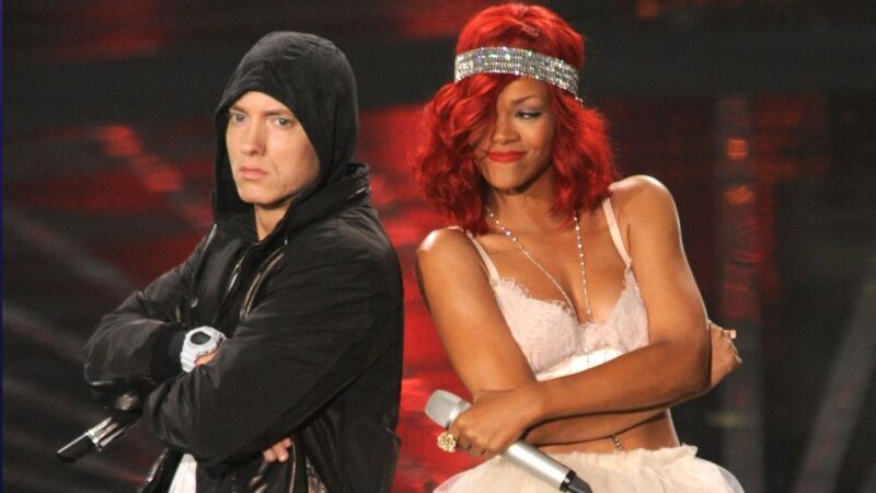Rihanna e Eminem em performance ao vivo, com os braços cruzados e expressão séria. Eminem está à esquerda usando um moletom preto com capuz na cabeça, enquanto Rihanna, à direita, tem cabelo vermelho curto, tiara brilhante prateada, top branco de renda e saia clara, segurando um microfone na mão direita. Fundo escuro com iluminação vermelha.