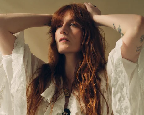 Fotografia de Florence. Na imagem, Florence Welch está com as mãos sobre a cabeça e usando um vestido branco. A imagem mostra a artista do peito para cima na frente de um plano de fundo bege e dá destaque para algumas tatuagens no braço esquerdo de Florence. O olhar da cantora é sereno e se direciona ao horizonte.