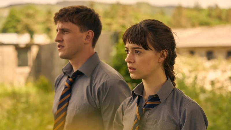 Cena da série Pessoas Normais. Na imagem, há um homem e uma mulher brancos de cabelos castanhos utilizando um uniforme cinza com uma gravata marcada por listras azuis e amarelas. Os dois estão em um jardim e olham para à frente.
