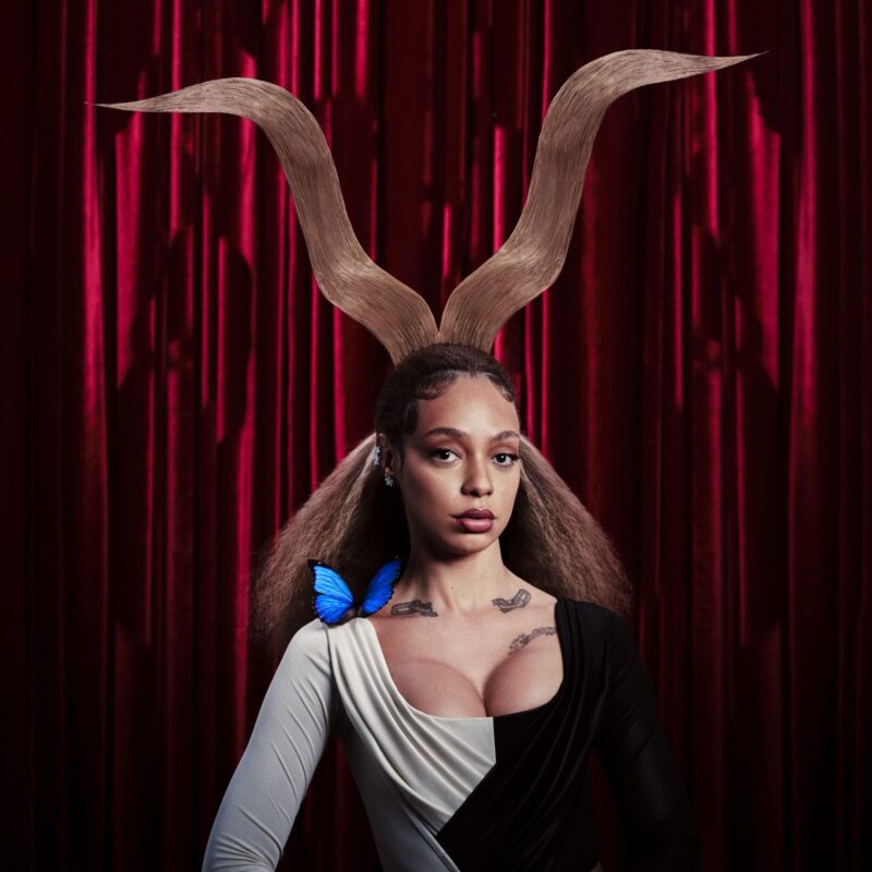 No centro da imagem, há uma mulher vestindo uma roupa preta e branca e com cabelos presos em um coque. Em sua cabeça, duas mechas de cabelo formam um chifre. Em seu ombro direito, há uma borboleta azul. 