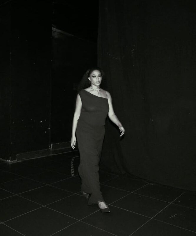 Na imagem, há uma mulher negra caminhando em um corredor escuro, vestindo um macacão de um ombro só. A foto está em preto e branco, e ao fundo é possível ver elementos de bastidores de um show.