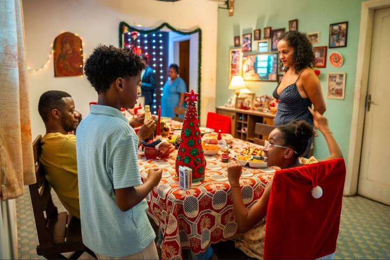 Uma família negra brasileira, composta por um homem sentado, uma mulher de pé e duas crianças, está reunida ao redor de uma mesa de jantar modesta. Sobre a toalha estampada vermelha, há pratos de comida e uma pequena árvore de Natal decorativa vermelha no centro. A mulher de pé olha para a criança sentada à direita.