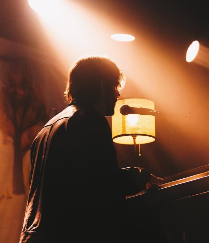 Um homem branco de cabelos castanhos curtos está sentado tocando piano e cantando. Não é possível ver seu rosto inteiro, apenas o perfil. Ele usa uma jaqueta escura que reflete a luz de um abajur amarelo e das luzes do palco ao fundo. Ele está inclinado para o microfone a sua frente. As luzes trazem um tom laranja e amarelo para a imagem. 