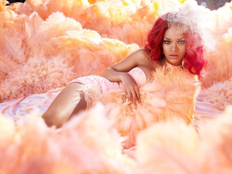 Rihanna na capa alternativa/foto promocional do álbum “Loud”: deitada de lado em um mar de plumas e pétalas em tons de pêssego e rosa suave, com cabelos vermelhos intensos no cabelo ondulado, véu de noiva delicado com rede na cabeça, vestido tomara-que-caia rosa claro drapeado, pele iluminada e expressão serena e confiante, olhando diretamente para a câmera. A imagem tem estética sonhadora, romântica e luxuosa.