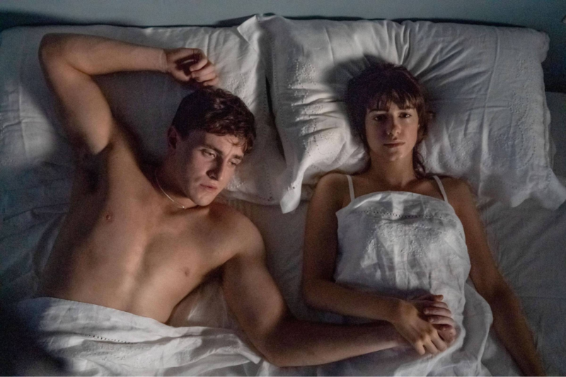 Cena da série Pessoas Normais. Na imagem, há um homem e uma mulher brancos de cabelos castanhos deitados em uma cama com cobertores, lençóis e travesseiros brancos. Eles estão de mãos dadas e sérios. Ele está sem blusa e ela está com um sutiã branco.