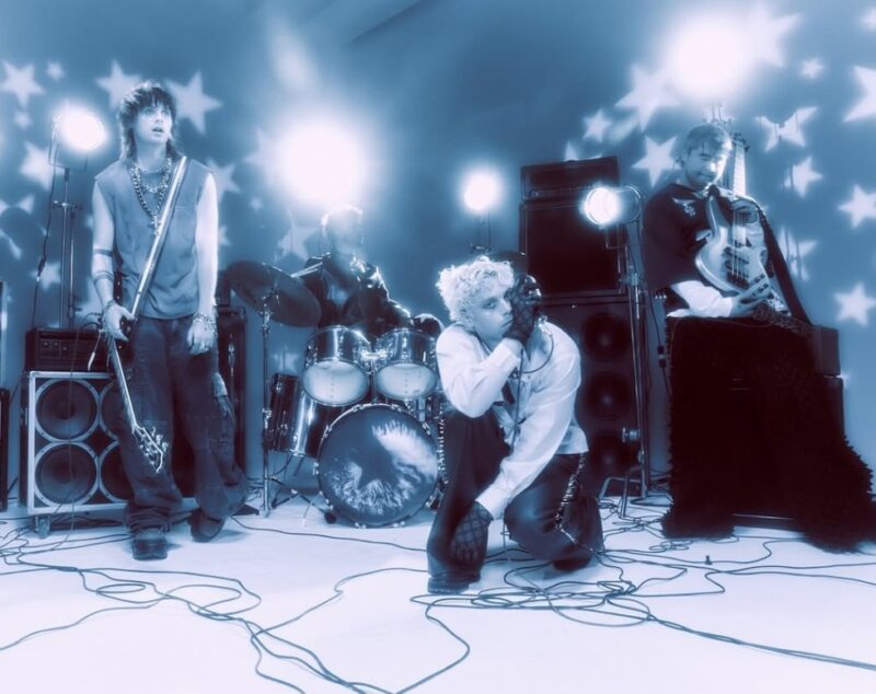 Quatro homens estão em um estúdio iluminado com luzes azuis que projetam estrelas brancas nas paredes. À esquerda, um deles segura a guitarra e tem a postura relaxada. Ao centro, um está posicionado atrás da bateria. Ao seu lado, levemente a frente, outro está agachado segurando um microfone. Ao lado direito, o outro está encostado em uma caixa de som e segurando um baixo. Atrás deles há caixas de som e de apoio dos instrumentos. No chão há cabos espalhados e conectando tudo.