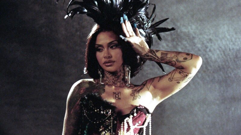 No centro da imagem, há uma mulher com tatuagens no pescoço, ombro e braços. Ela está com a mão levantada na altura do rosto enquanto veste um espartilho com detalhes de pérolas, brincos grandes e cabelo curto. Ela também possui um adorno de penas na cabeça.