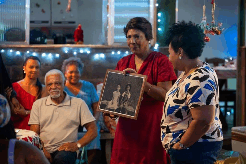 Uma mulher negra mais velha, usando um vestido vermelho, segura e exibe um quadro com uma fotografia antiga em preto e branco de duas meninas. Ela sorri enquanto outra mulher ao seu lado olha para a foto. Ao fundo, um homem idoso de camisa polo cinza está sentado, observando a cena. O ambiente é um pátio iluminado por luzes de Natal.