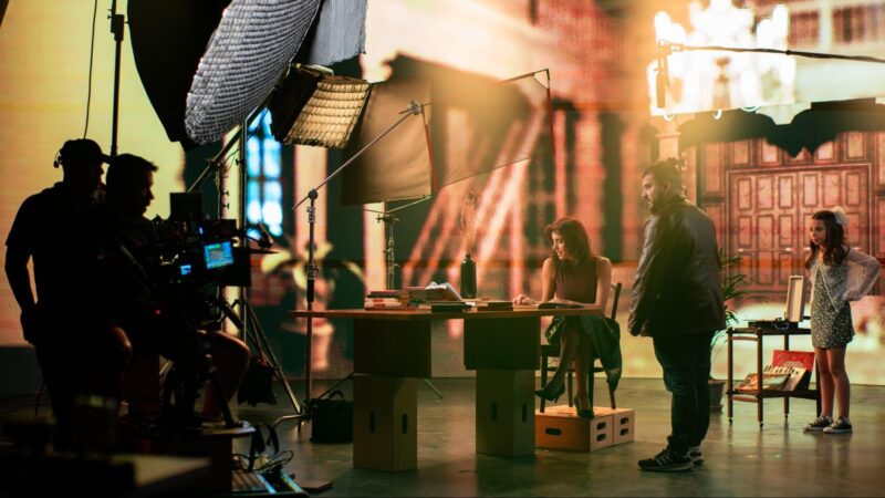 A foto apresenta um estúdio de gravação cinematográfica iluminado por luzes quentes e preenchido por técnicos e equipamentos que cercam uma mesa onde uma mulher morena sentada consulta algo. Próximo a ela, um homem de coque e jaqueta de couro observa a cena enquanto uma menina também morena, ao fundo, permanece em pé com expressão curiosa. 