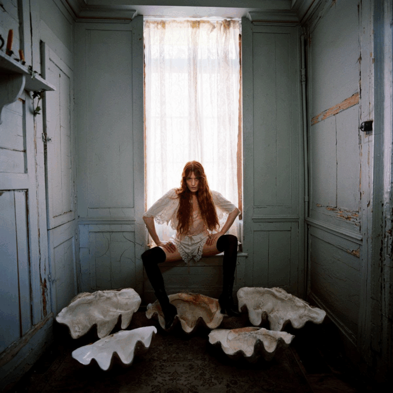 Fotografia de Florence. Na imagem, Florence Welch está sentada contra a luz de pernas abertas na janela de um corredor de aparência antiga e sombria. A artista veste um conjunto branco similar a um pijama. No chão, próximo às suas pernas, alguns vasos de cerâmica com bordas onduladas. As paredes de madeira ao redor estão descascadas em algumas extremidades.