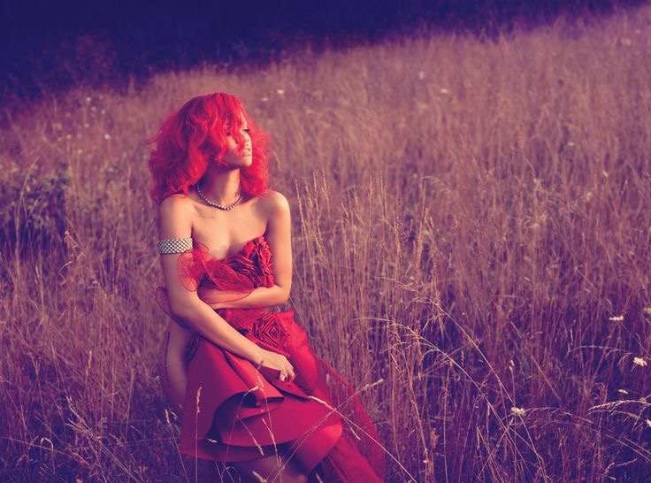 Rihanna de cabelos vermelhos vibrantes está sentada em um campo de grama alta e seca ao entardecer. Ela usa um vestido vermelho estruturado com detalhes florais no busto, um colar de pérolas e uma pulseira prateada no braço. A iluminação é suave e quente, com tons avermelhados dominando a cena, criando uma atmosfera etérea e romântica. Ela olha para o lado com expressão serena.