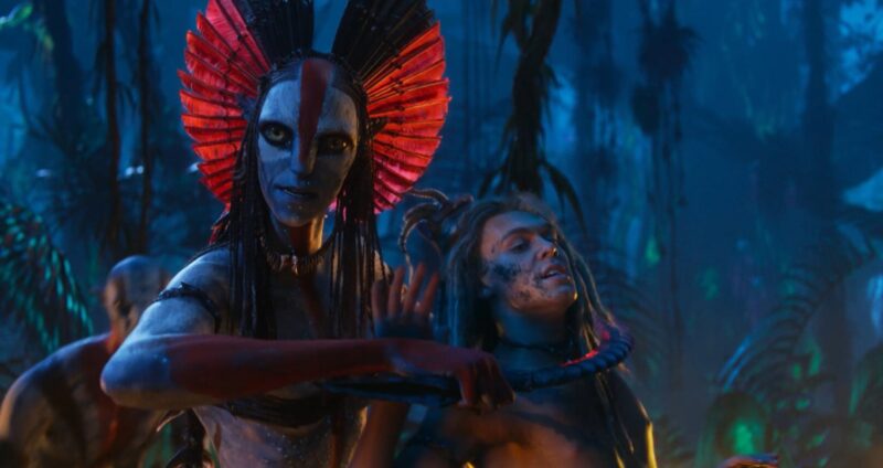 Cena do filme Avatar: Fogo e Cinzas. Na imagem, Varang mantém Spider como refém. Ela aponta uma lâmina para o pescoço do rapaz com uma mão enquanto segura os cabelos dele com a outra. Ela é uma alienígena, de pele azul, adorno vermelho de penas na cabeça, tintura branca e vermelha no rosto e braços. Spider é um garoto na faixa dos 20 anos, de pele clara e cabelos loiros trançados. Eles estão na floresta, é noite e em volta deles há outros aliens. 