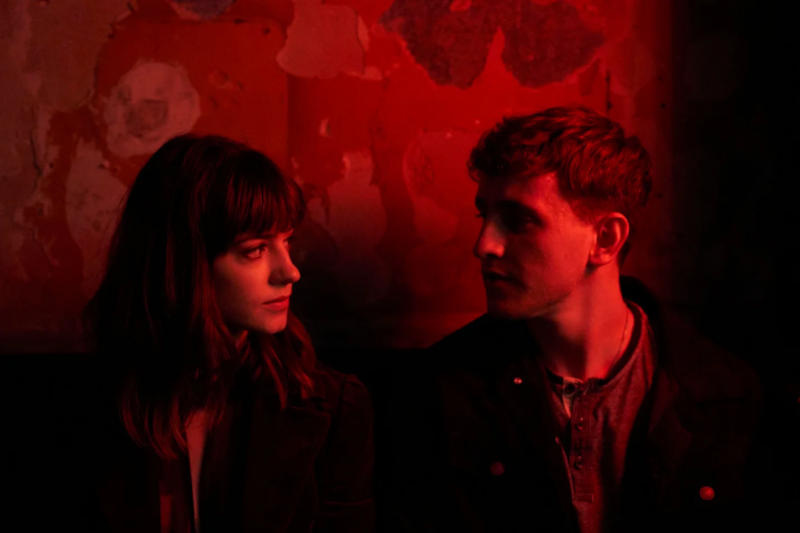 Cena da série Pessoas Normais. Na imagem, há uma mulher branca de cabelos castanhos e franja olhando para um homem branco de cabelos castanhos, que também devolve o olhar. Eles estão sentados e uma luz vermelha ilumina o espaço onde estão.