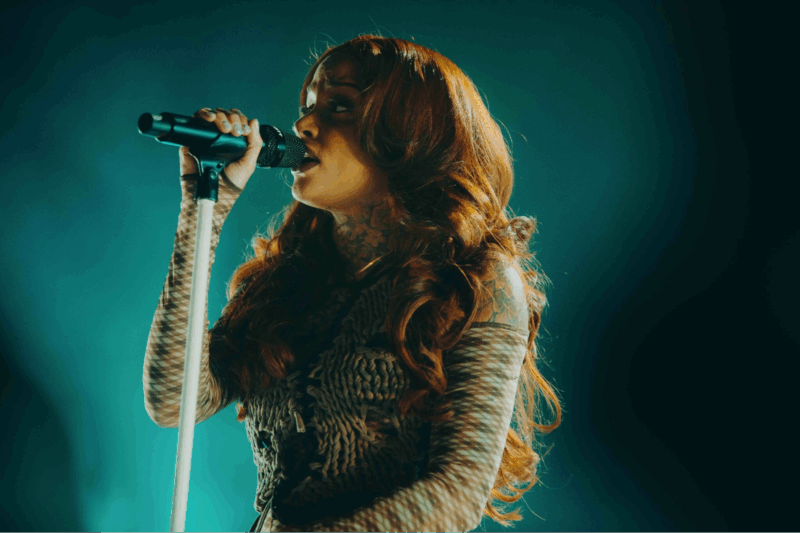 No centro da imagem, há uma mulher cantando enquanto segura o microfone. Ela está vestindo uma blusa e usando luvas que cobrem os braços, com o cabelo longo e ondulado solto. O fundo da imagem é predominantemente azul.