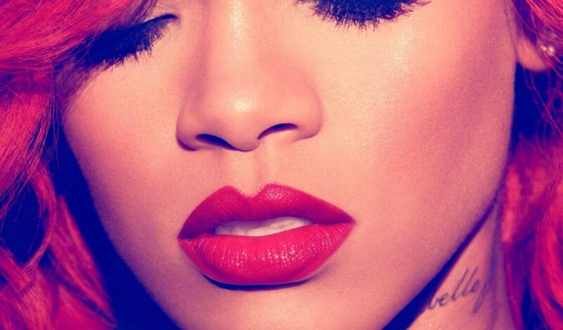 Close-up da capa do álbum “Loud” de Rihanna: rosto da cantora em tons quentes de vermelho e rosa, com cabelos ruivos volumosos caindo sobre os ombros, olhos fechados maquiados com sombra preta esfumada, batom vermelho intenso e brilhante nos lábios entreabertos, e uma tatuagem com a palavra “rebelle” visível no pescoço. A imagem transmite sensualidade e atitude marcante.