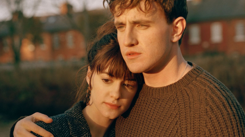 Cena da série Pessoas Normais. Na imagem, há uma mulher e um homem brancos de cabelos castanhos abraçados. Ela usa uma blusa preta e ele veste um suéter marrom. Os dois estão sérios e acompanhados do sol, que reflete em seus rostos.