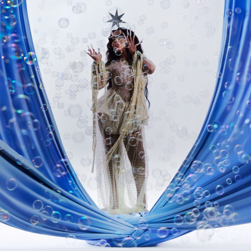 No centro da imagem, há uma mulher com vestido prateado e com cabelos escuros e longos. Ela está com as mão no rosto e possui unhas longas e um adorno em formato de estrela na cabeça. Ao seu redor, há elementos em azul e, por toda a imagem, há bolhas de sabão. 