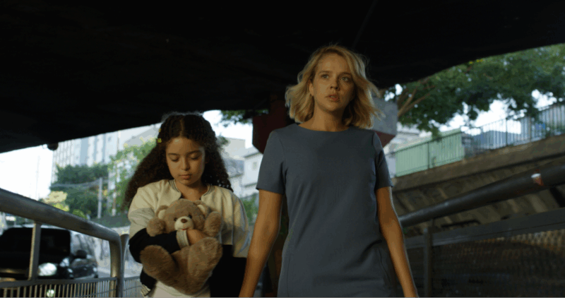 Em um recorte do filme, uma mulher loira de olhos claros de vestido azul caminha à frente com expressão apreensiva olhando à sua frente, enquanto uma menina morena com presilhas no cabelo ao seu lado segura um urso de pelúcia bege, olhando para baixo. Ao fundo, uma passagem urbana sob um viaduto. 