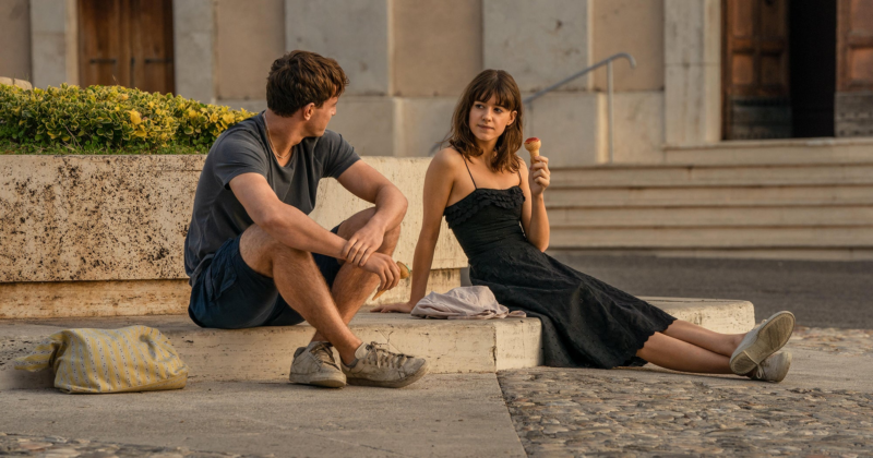 Cena da série Pessoas Normais. Na imagem, há um homem e uma mulher brancos de cabelos castanhos sentados na calçada. Ela usa um vestido preto e um tênis branco, além de segurar um sorvete na mão esquerda. Ele veste uma blusa e short azuis, além de usar um tênis branco. Os dois se encaram.