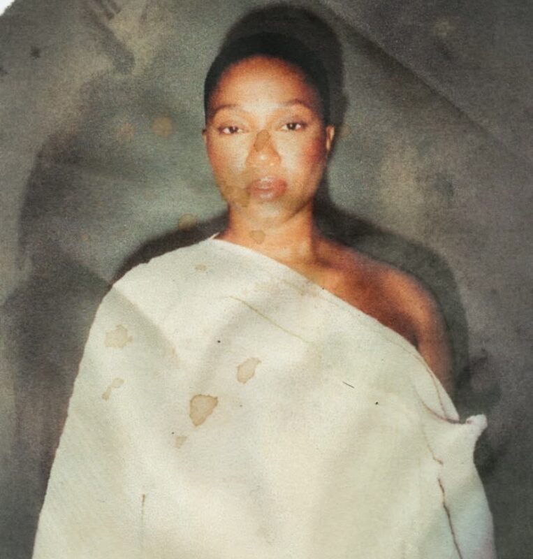 Na imagem, há o retrato de uma mulher com expressão serena, olhando diretamente para a câmera. Ela está vestida com tecidos em cores claras, que cobre um dos ombros. A foto tem textura envelhecida, com manchas e marcas que lembram desgaste de papel antigo.