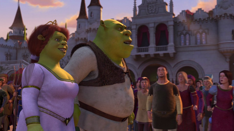 Frame de Shrek, ogro verde com roupa marrom e branca, e Fiona, ogra verde de cabelo vermelho e vestido roxo e detalhes dourados, se aproximando da entrada de um palácio, um castelo branco com vários andares. Enquanto eles caminham, as pessoas ao seu redor observam chocadas com a aparência dos dois.