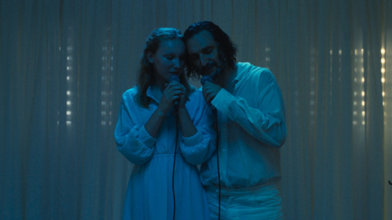 Sob uma iluminação fria e azulada, Rebecca (Helena Zengel) e seu pai Lawrence (Jeremy Xido) estão de pé, lado a lado, segurando microfones próximos à boca. Ambos vestem roupas brancas e parecem cantar ou orar de olhos fechados ou baixos. O fundo é composto por uma cortina branca translúcida.