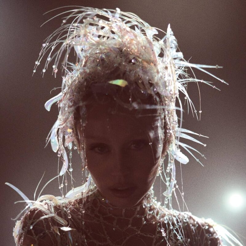 Texto Alt: Miley Cyrus aparece de frente, com o rosto envolto por fios translúcidos e brilhantes, iluminados por uma luz forte ao fundo. O fundo é escuro, criando um efeito dramático e etéreo. Flávia Ferracini