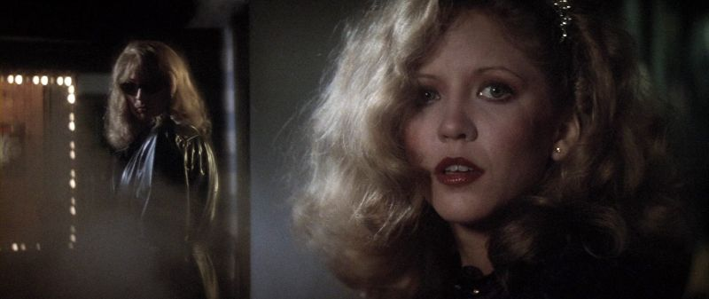 Cena de Vestida para Matar. No primeiro plano, à direita, Liz olha para o lado com expressão de apreensão ou surpresa. Ao fundo, à esquerda, uma figura misteriosa com óculos escuros e casaco brilhante a observa das sombras em um ambiente mal iluminado com luzes desfocadas.