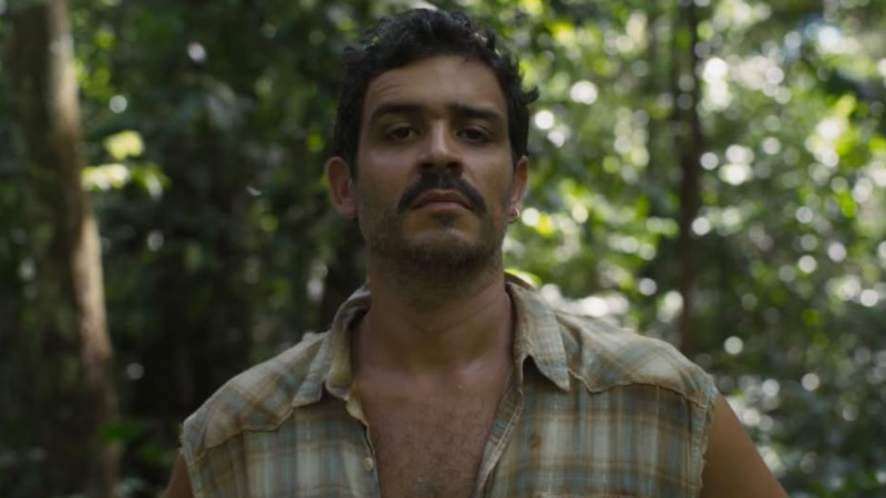 Close de Philipp Lavra de frente para a câmera em meio a uma floresta. Ele tem cabelos escuros, bigode e aparenta estar suado. Usa uma camisa xadrez bege sem mangas com os botões abertos. O fundo é um desfoque de folhagens verdes e luz solar filtrada.