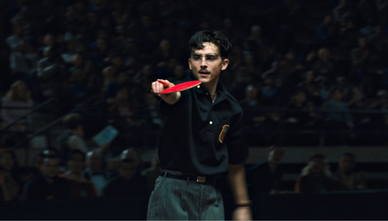 Cena de Marty Supreme. O cenário é de um ginásio com iluminação dramática. O ator Timothée Chalamet, caracterizado com bigode e óculos de grau, está em foco no centro da imagem. Ele veste uma camisa polo preta com um escudo bordado no peito e calças sociais cinzas. Ele segura uma raquete de tênis de mesa vermelha, apontando-a para a frente em uma pose de ação. O fundo está desfocado, mostrando uma plateia em uma arena esportiva escurecida.