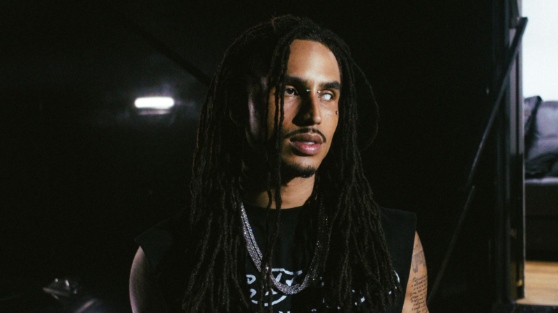 A imagem mostra um homem negro de cabelos longos em dreadlocks, iluminado por luz dura em um ambiente escuro de bastidores. O olhar se projeta para fora do quadro, distante e atento, como se carregasse o peso do silêncio entre o palco e a solidão.