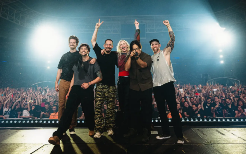 Seis músicos reunidos no centro do palco ao final de um show, de braços levantados e sorrindo, diante de uma multidão com as mãos erguidas. Luzes fortes iluminam a cena, destacando o entusiasmo coletivo entre artistas e público.