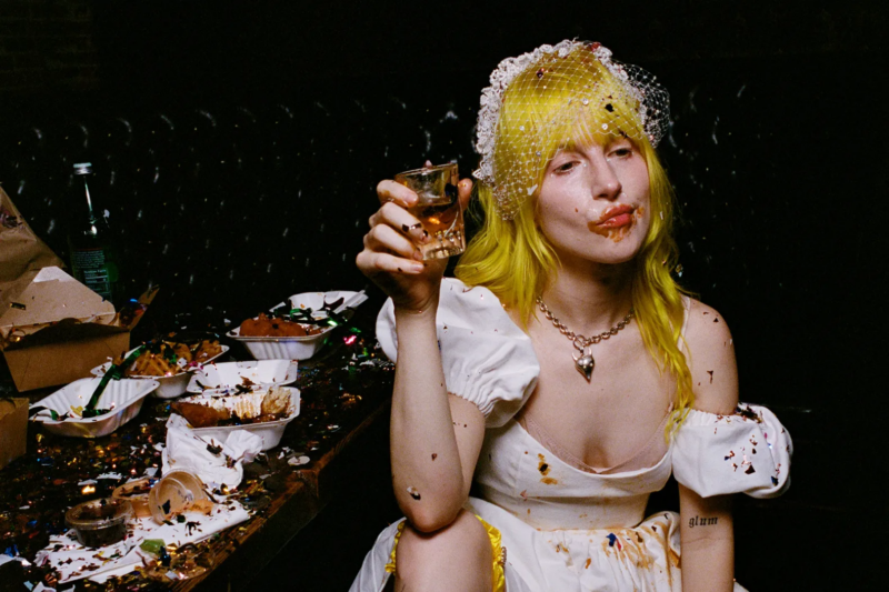 Texto Alternativo: Fotografia de Hayley Williams. Na imagem, a cantora aparece sentada, com um vestido e um véu de noiva sujos de comida. A boca de Williams também está suja, ela parece mastigar algo. Ela segura um copo de bebida cheio até a metade. À esquerda, há uma mesa com embalagens de comida reviradas e espalhadas.