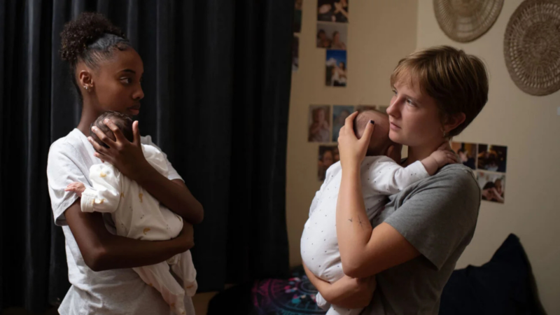Em um quarto com pouca luz, duas jovens mulheres estão de pé, de frente uma para a outra, cada uma segurando um bebê pequeno apoiado no ombro. A jovem da esquerda é negra e veste uma camiseta branca; a da direita tem cabelo loiro curto e veste uma camiseta cinza. Elas se olham nos olhos com expressões sérias e cansadas. Ao fundo, uma parede com fotos coladas e uma cortina escura.