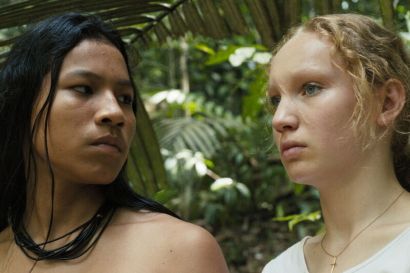Um close-up na floresta mostra Silas (Hamã Sateré), um jovem indígena de cabelos escuros e colar de contas olhando seriamente para a esquerda, ao lado de Rebecca (Helena Zengel), uma jovem branca de cabelos loiros cacheados olhando para a direita. Ela usa um colar com uma pequena cruz dourada. O fundo é uma vegetação densa e desfocada.