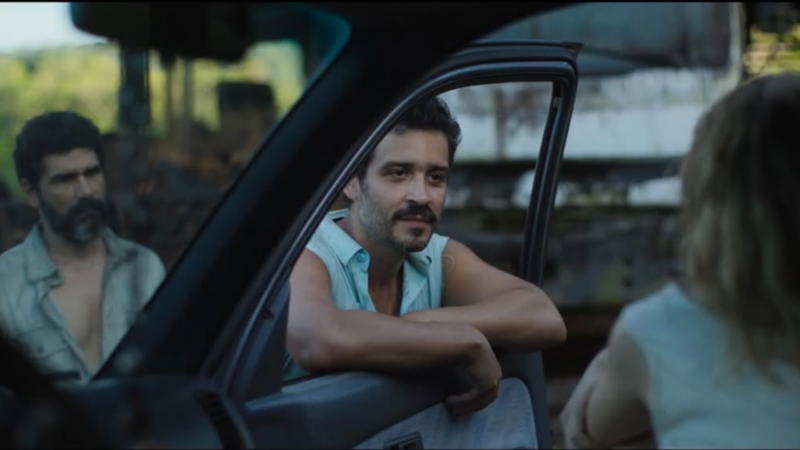 Plano médio mostra Philipp Lavra debruçado na janela de um carro, olhando para a direita. Ele usa uma camisa azul clara sem mangas e tem bigode. Ao fundo, fora de foco, o ator Rômulo Braga observa a cena com expressão séria.