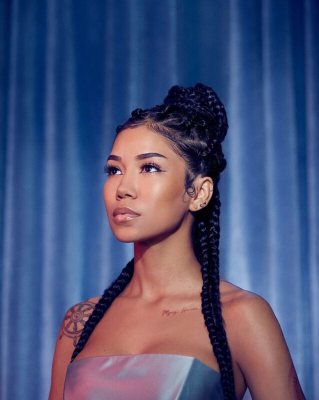 Na imagem, há um retrato de uma mulher com tranças longas presas em um penteado alto. Ela usa um top metálico prateado e tem tatuagens visíveis no ombro e na clavícula. O fundo é azul e desfocado, dando destaque ao rosto e à iluminação suave. 