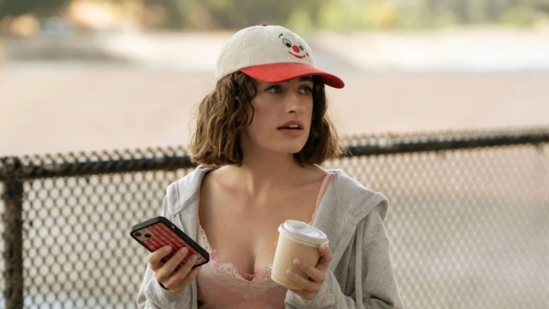 Cena da série I Love LA. Na imagem, há uma mulher branca de cabelos ondulados que veste uma blusa rosa com uma jaqueta cinza, além de utilizar um boné branco e vermelho com estampa de um palhaço. Ela está em pé e segura um celular na mão direita e um café na mão esquerda.