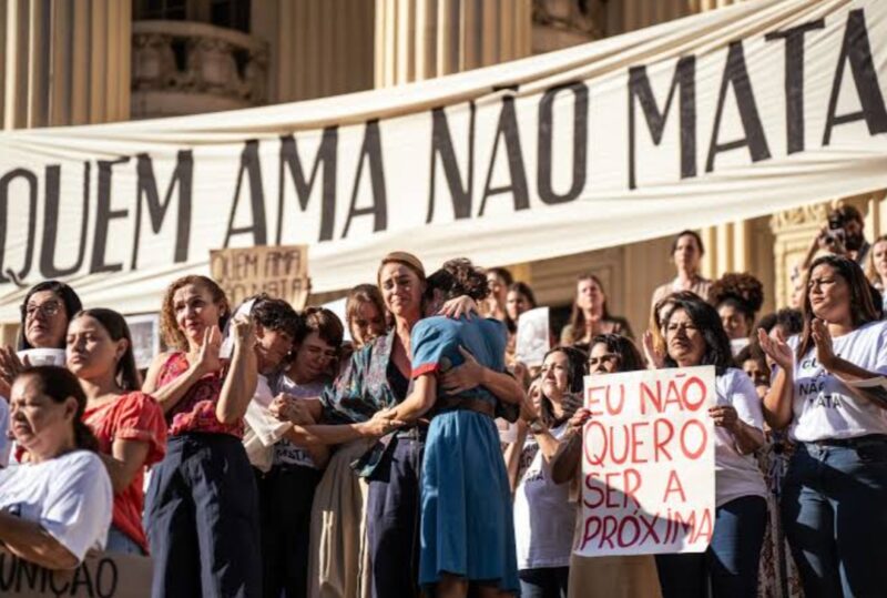 Cena de Ângela Diniz: Assassinada e Condenada. Grupo de mulheres reunidas em protesto em frente a um prédio com colunas. Ao fundo, uma grande faixa com os dizeres "Quem ama não mata". No centro, uma mulher abraça outra emocionada. Algumas manifestantes aplaudem e uma segura um cartaz escrito ‘Eu não quero ser a próxima’ em vermelho.
