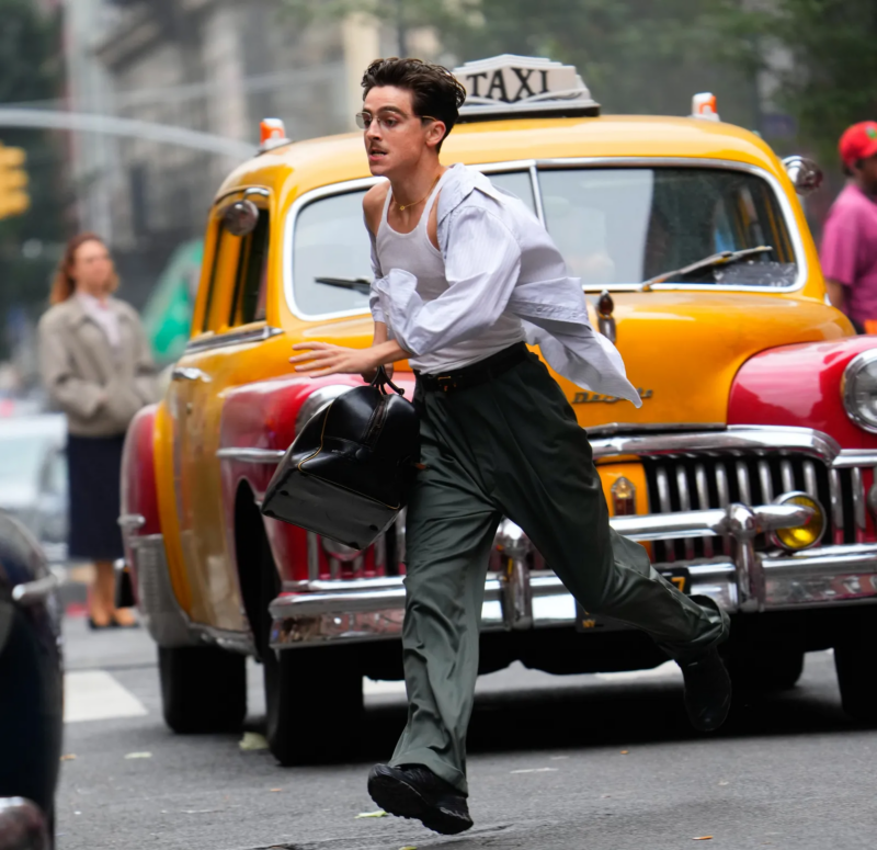 Cena de Marty Supreme. Cenário é uma rua urbana movimentada. Timothée Chalamet é capturado em pleno movimento, correndo rapidamente para o lado esquerdo da imagem. Ele carrega uma bolsa de couro preta e veste uma regata branca por baixo de uma camisa social clara aberta e desabotoada, que voa com o vento. Ao fundo, um táxi clássico amarelo e vermelho